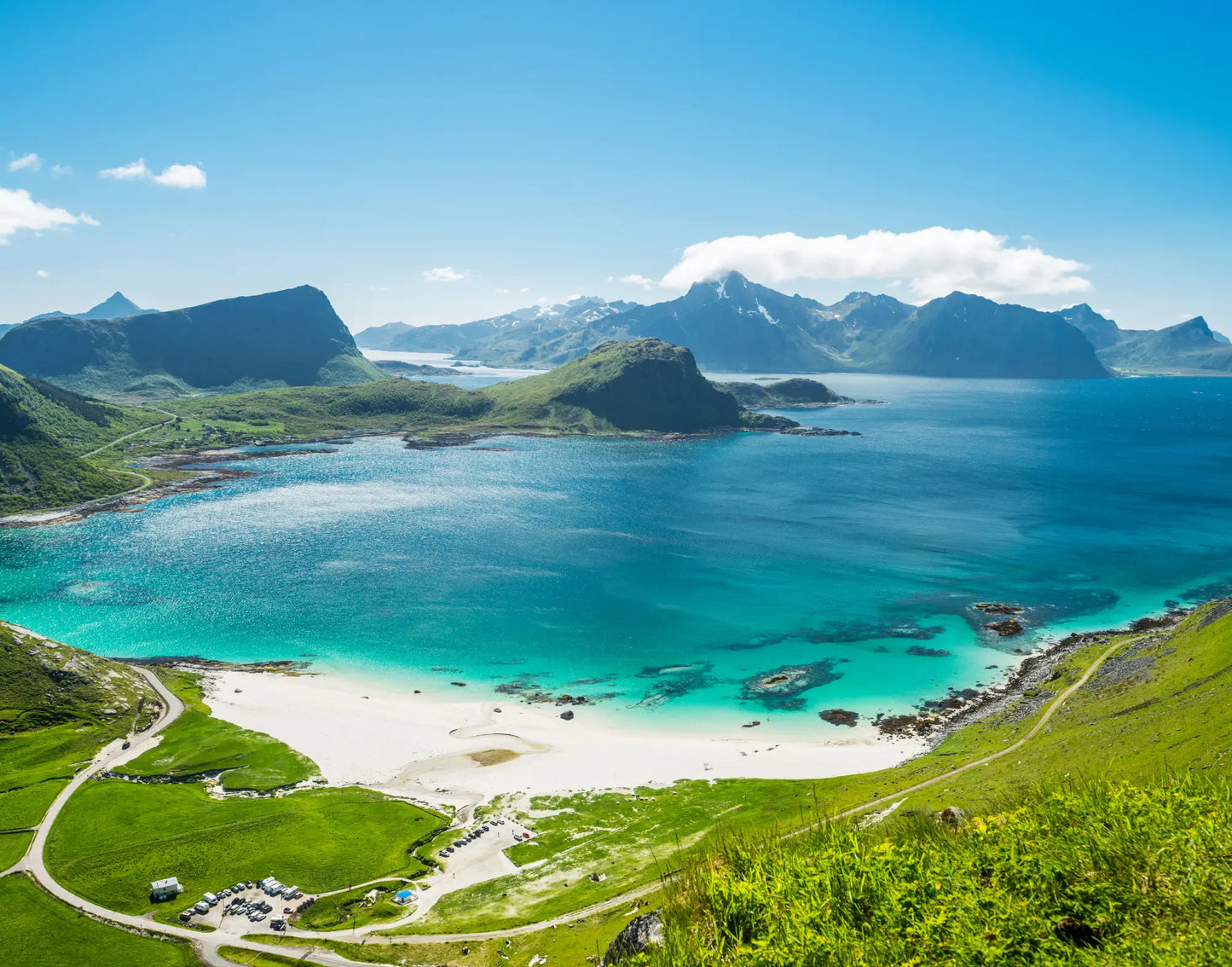 Haukland beach in Lofoten, Norway. The white sandy beach has beautiful turquoise water, and is surrounded by mountain peaks. It’s a summer day, the mountains are covered in green grass, sun is shining from a clear blue sky.