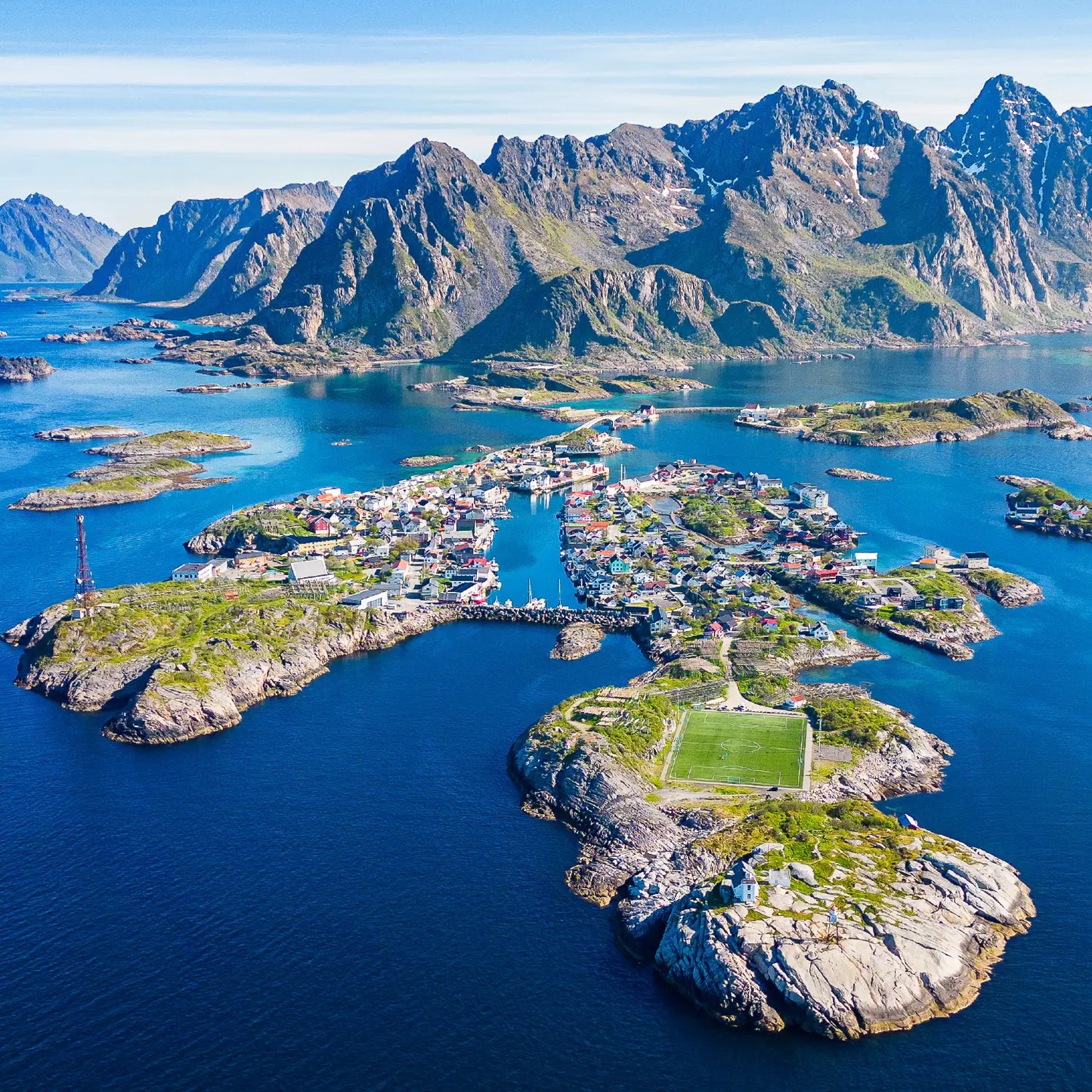 Henningsvær in Lofoten seen from above. Beautiful blue sky, mountain peaks. The sun is shining on the football field. The ocean is blue
