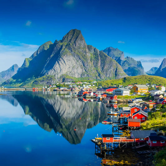 Traditional red fisherman cabins in front of a big beautiful mountain in Reine, Lofoten. The sun is shining and the water is blue.